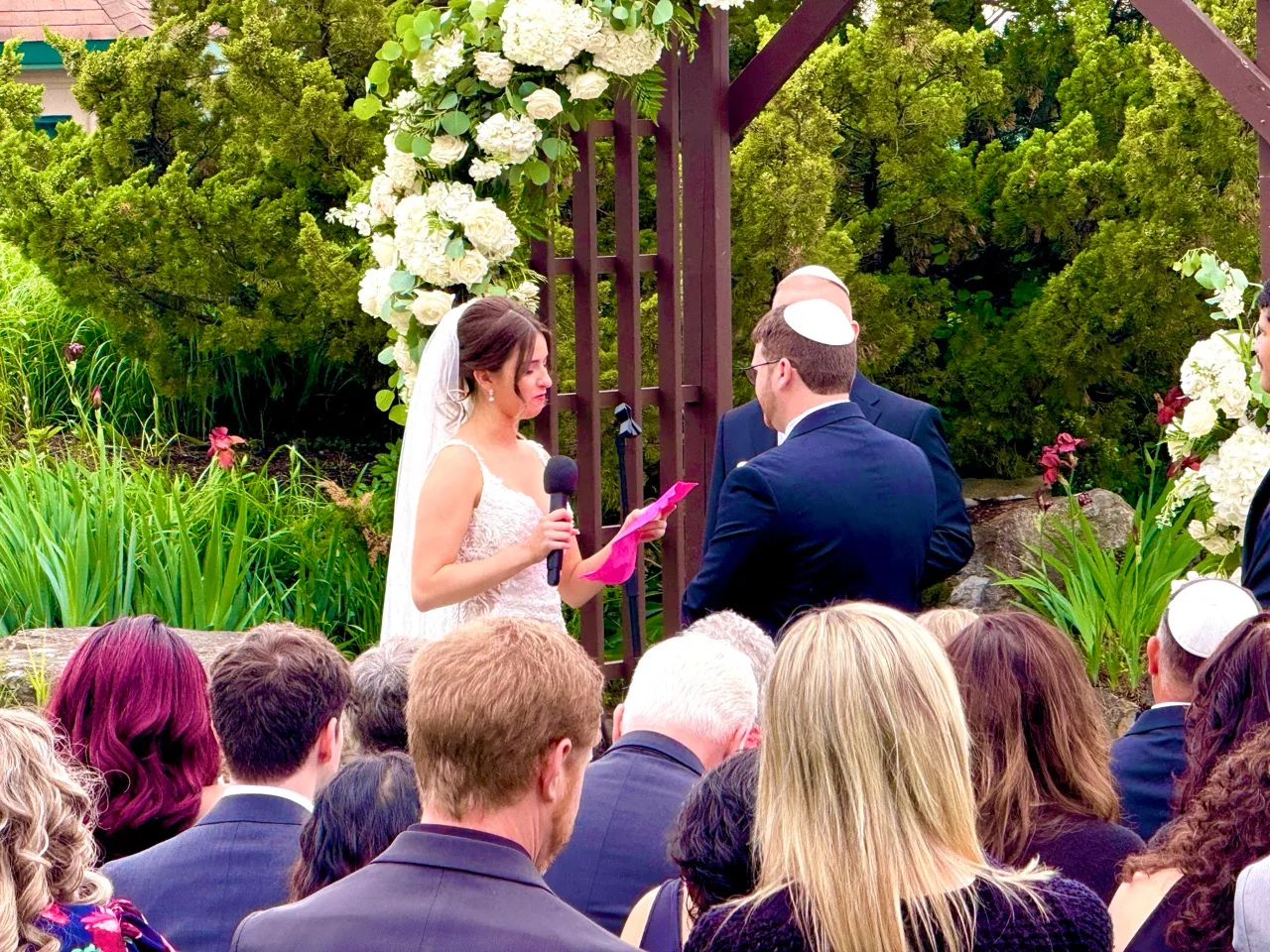 Bride reading her vows into a handheld microphone beneath a floral ceremony arch