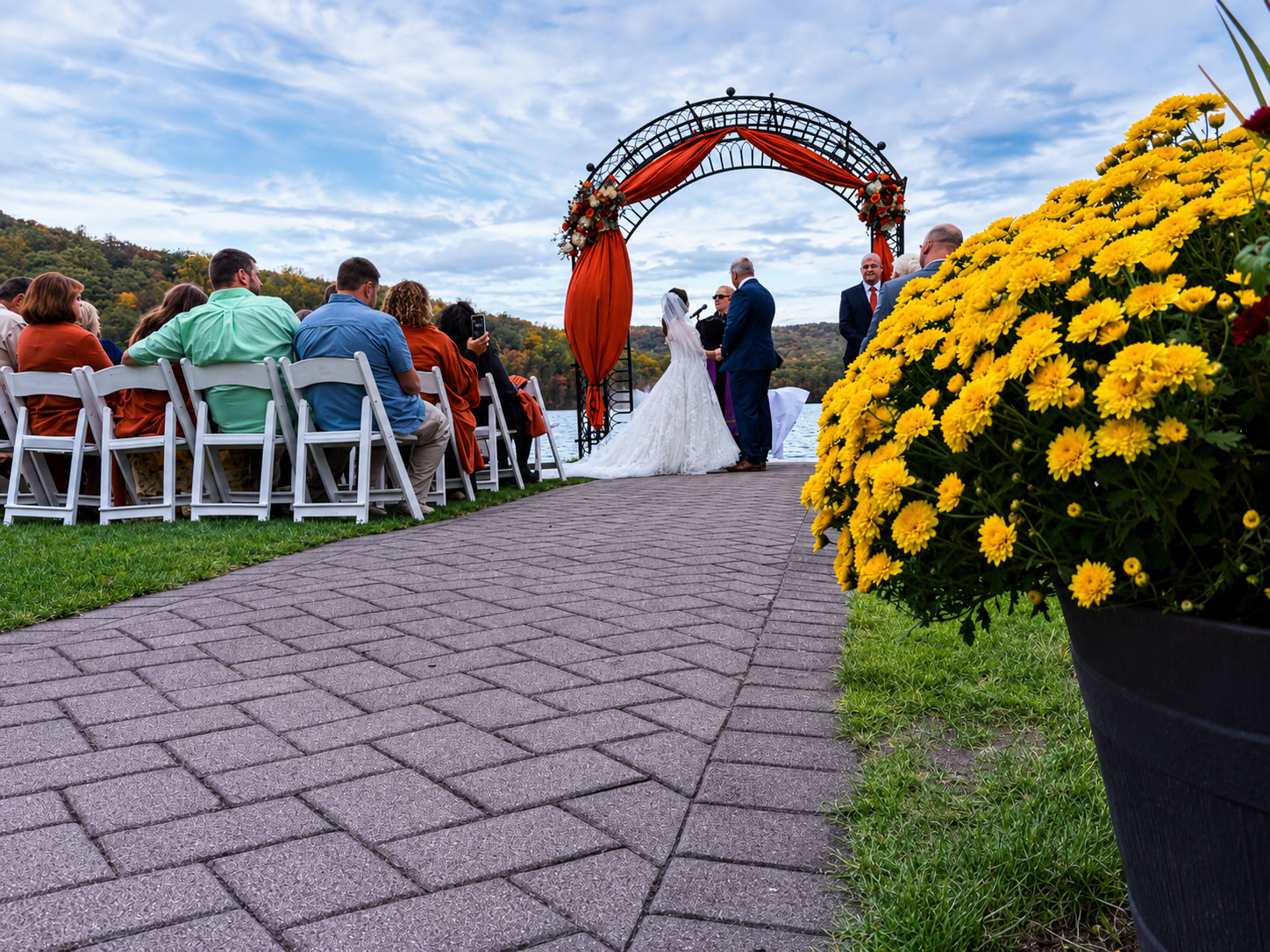 Lakeside wedding ceremony at Lake Valhalla Club with an orange-draped iron arch and autumn mums beside the water